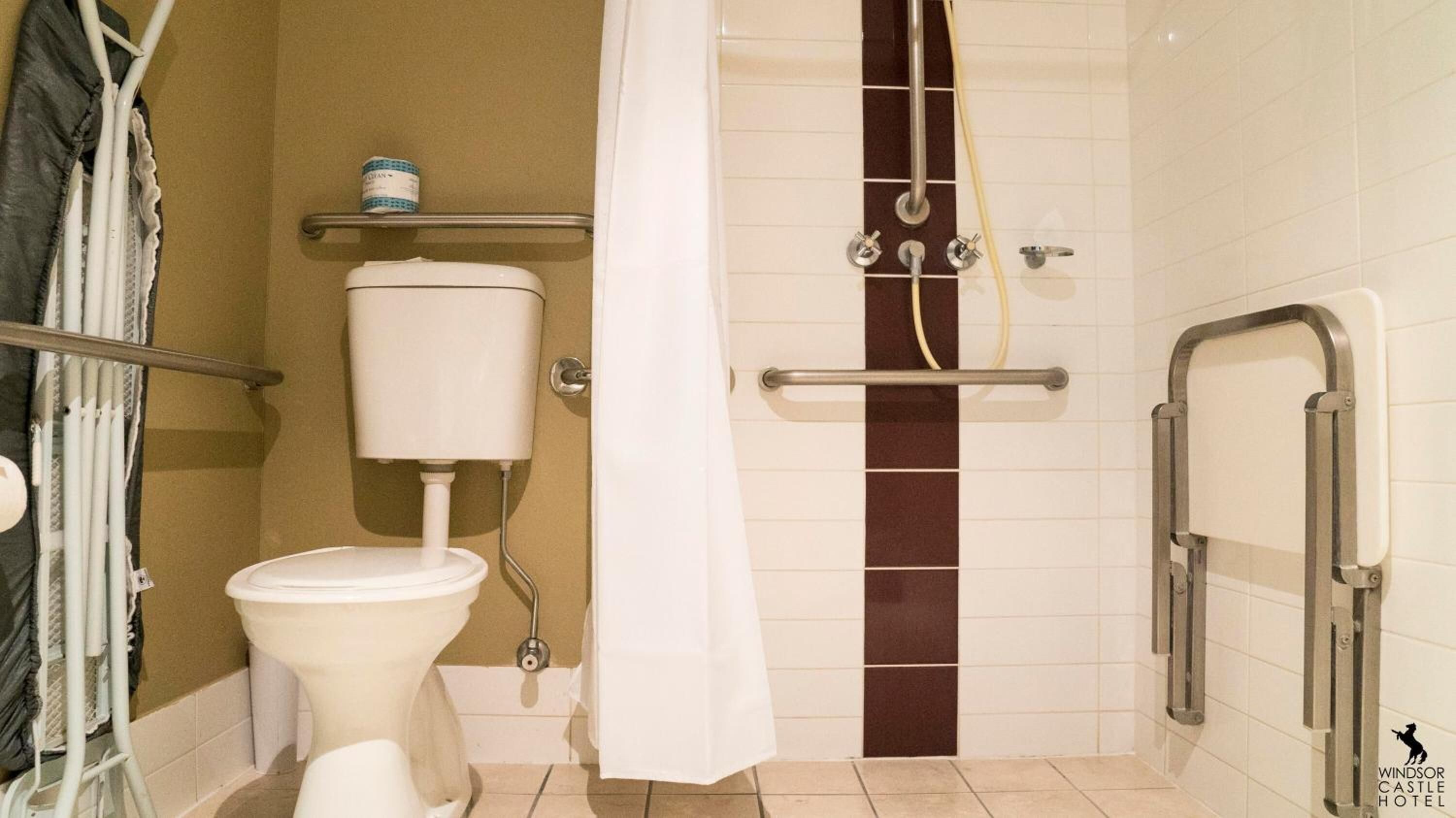 Un baño con inodoro, ducha con barras de apoyo, toallero y estante con artículos de aseo.