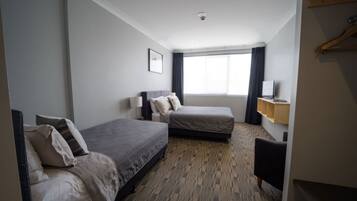 Quarto de hotel com duas camas de solteiro, uma escrivaninha, uma cadeira, uma televisão e uma janela com cortinas.