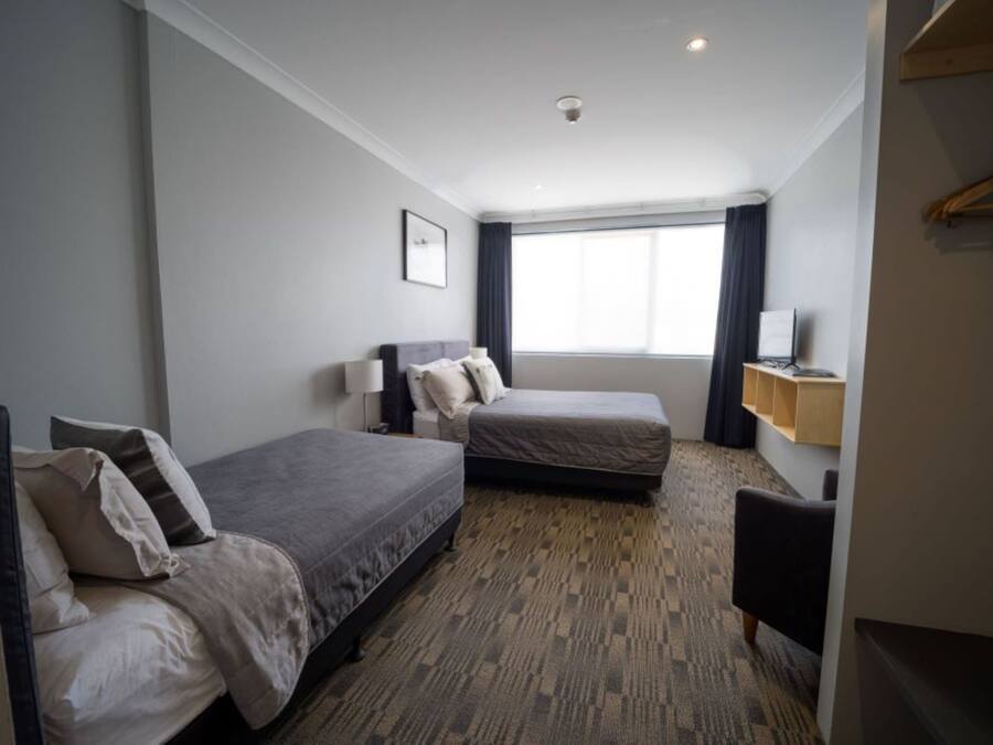 Quarto de hotel com duas camas de solteiro, uma escrivaninha, uma cadeira, uma televisão e uma janela com cortinas.