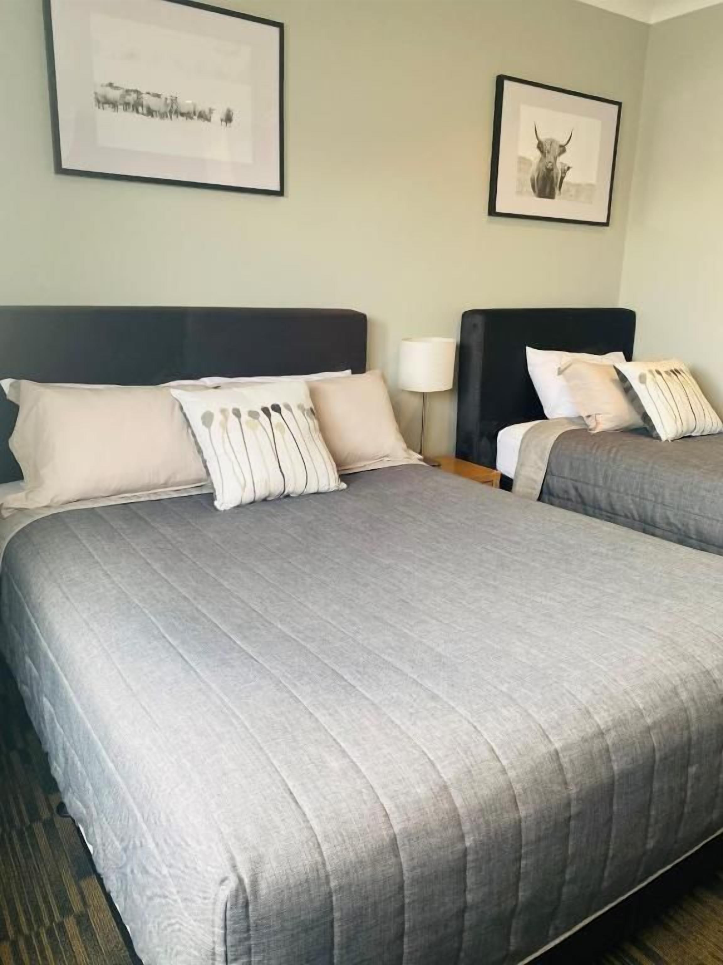 Dos camas con ropa de cama gris, una mesita de noche con lámpara y cuadros en las paredes.