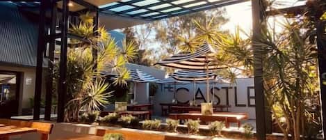 Un área de comedor al aire libre con mesas y bancos de madera, techo de vidrio y vista a un edificio que dice 'THE CASTLE'.