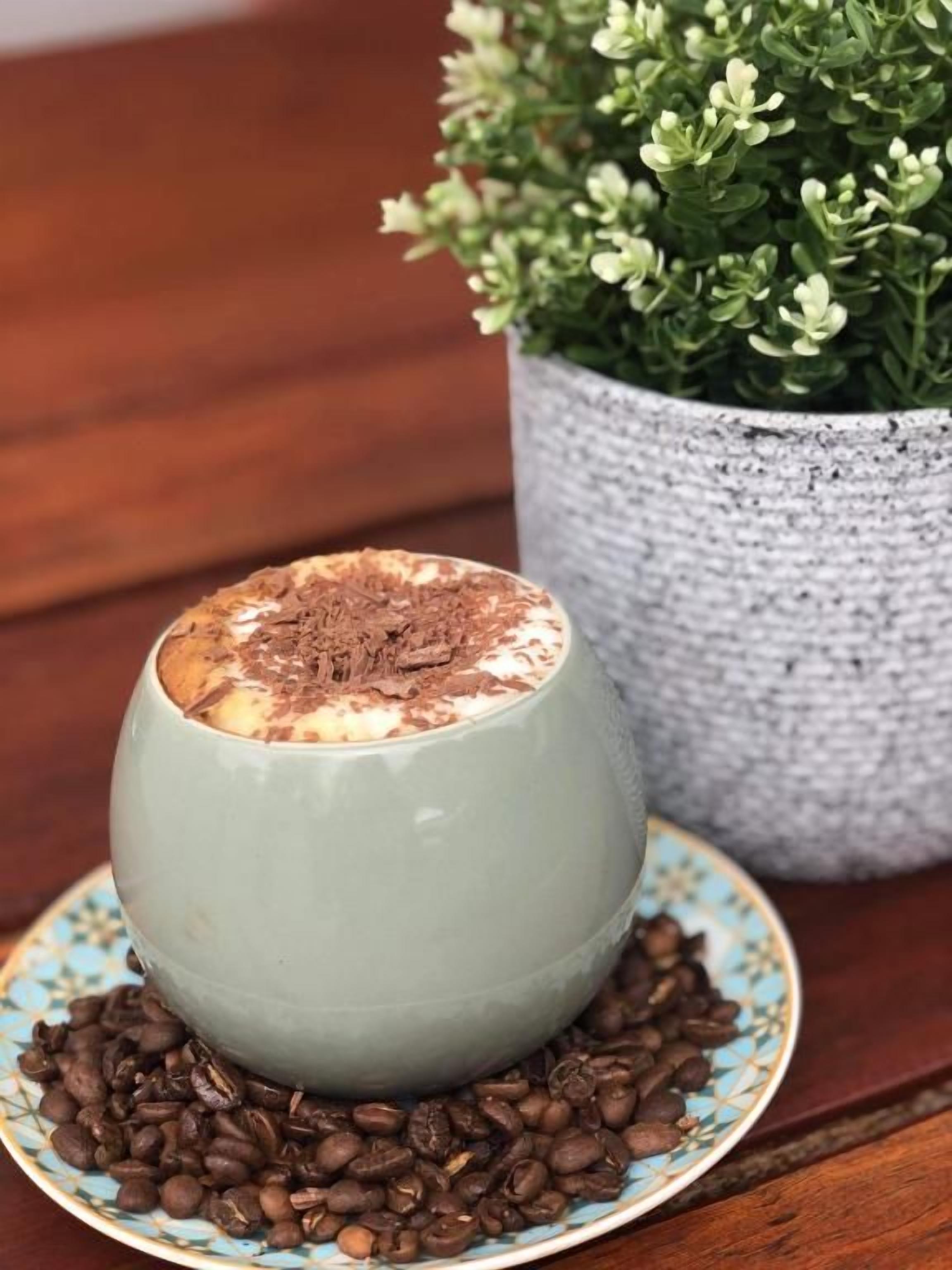 A ceramic mug with a chocolate top, placed on a plate surrounded by coffee beans, with a potted plant in the background.