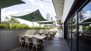 Un patio exterior con mesas y sillas bajo sombrillas verdes, piso de concreto y vista de la vegetación circundante.