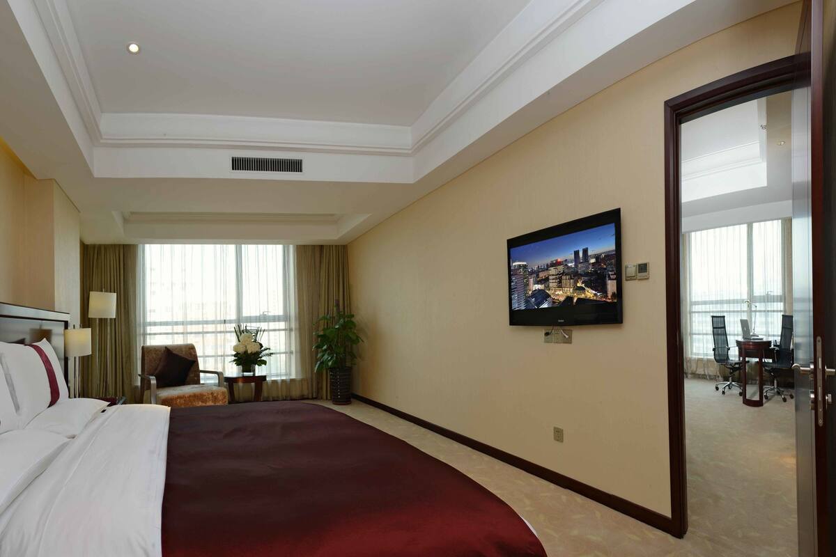 Ein Hotelzimmer mit einem großen Bett, einem Fernseher, einem Schreibtisch mit zwei Stühlen und Blick auf die Stadt durch das Fenster.