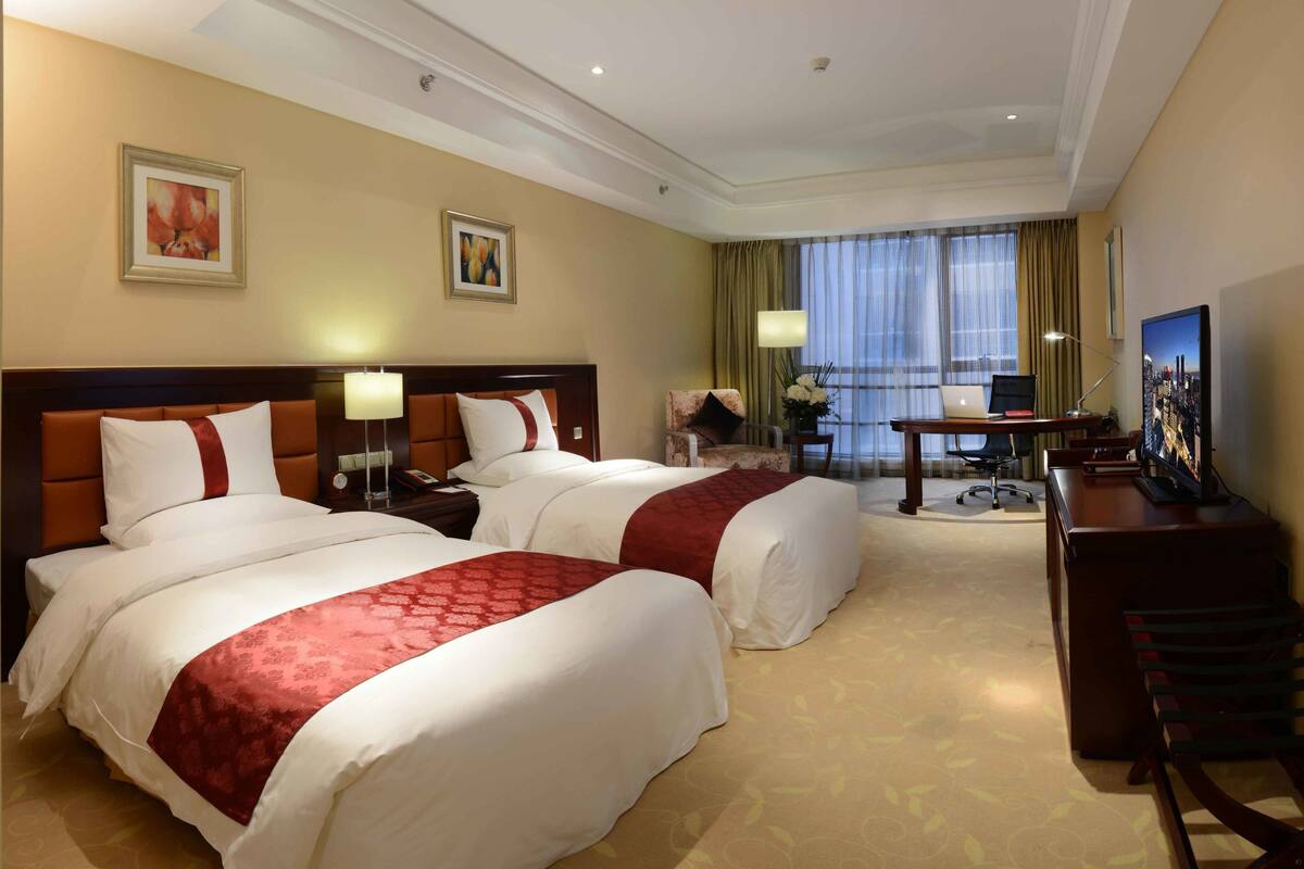 Ein Hotelzimmer mit zwei Betten, einem Schreibtisch, einem Stuhl, einem Fernseher und einem Fenster mit Vorhängen.