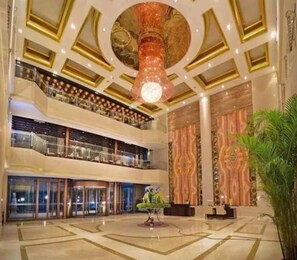 A multi-level hotel lobby with a grand chandelier, a reception desk, and a large plant.