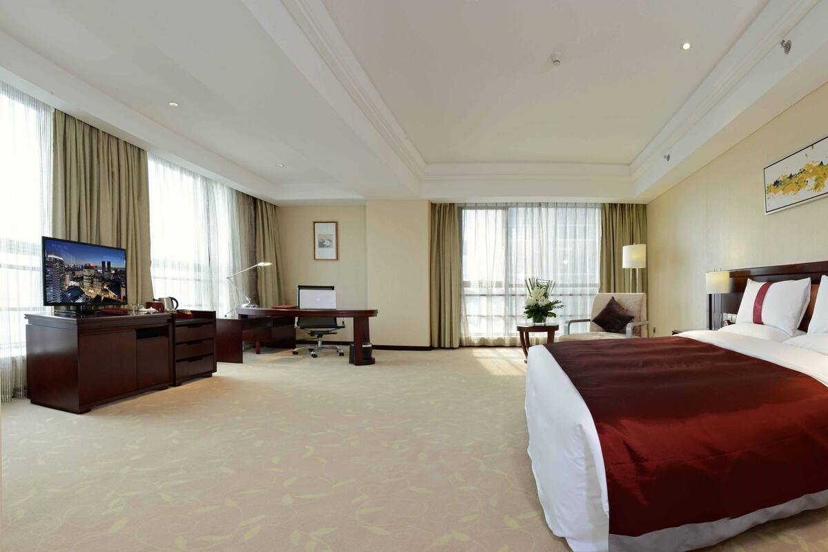 Ein Hotelzimmer mit einem großen Bett, einem Schreibtisch mit Computer, einem Fernseher und einem Fenster mit Vorhängen.