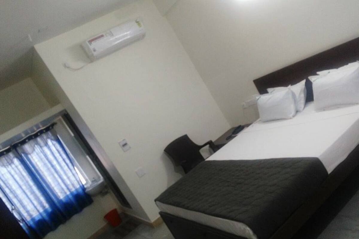 A hotel room with a bed, a bedside table, a chair, a window with curtains, and a wall-mounted air conditioner.