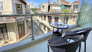 Un balcón con sillas de mimbre y una pequeña mesa, con vista a una calle con edificios y una torre de comunicaciones alta.