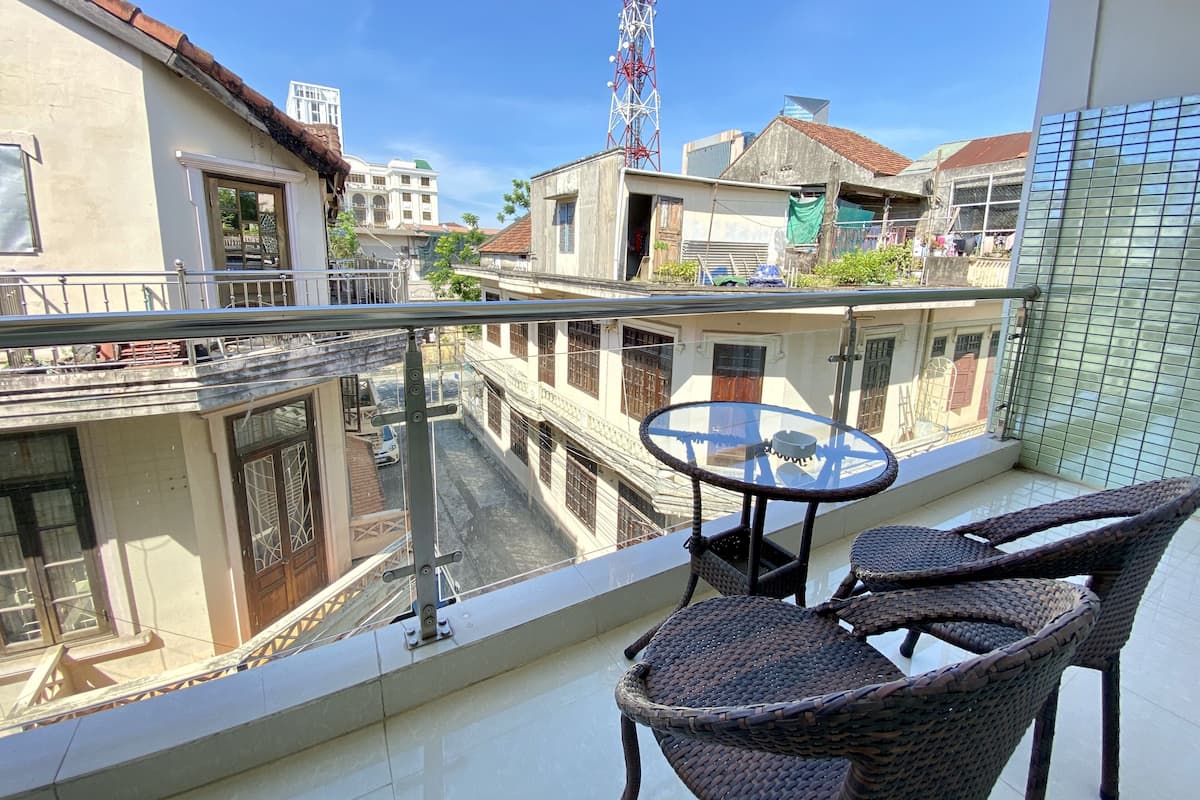 A balcony with wicker chairs and a small table, overlooking a street with buildings and a tall communication tower.