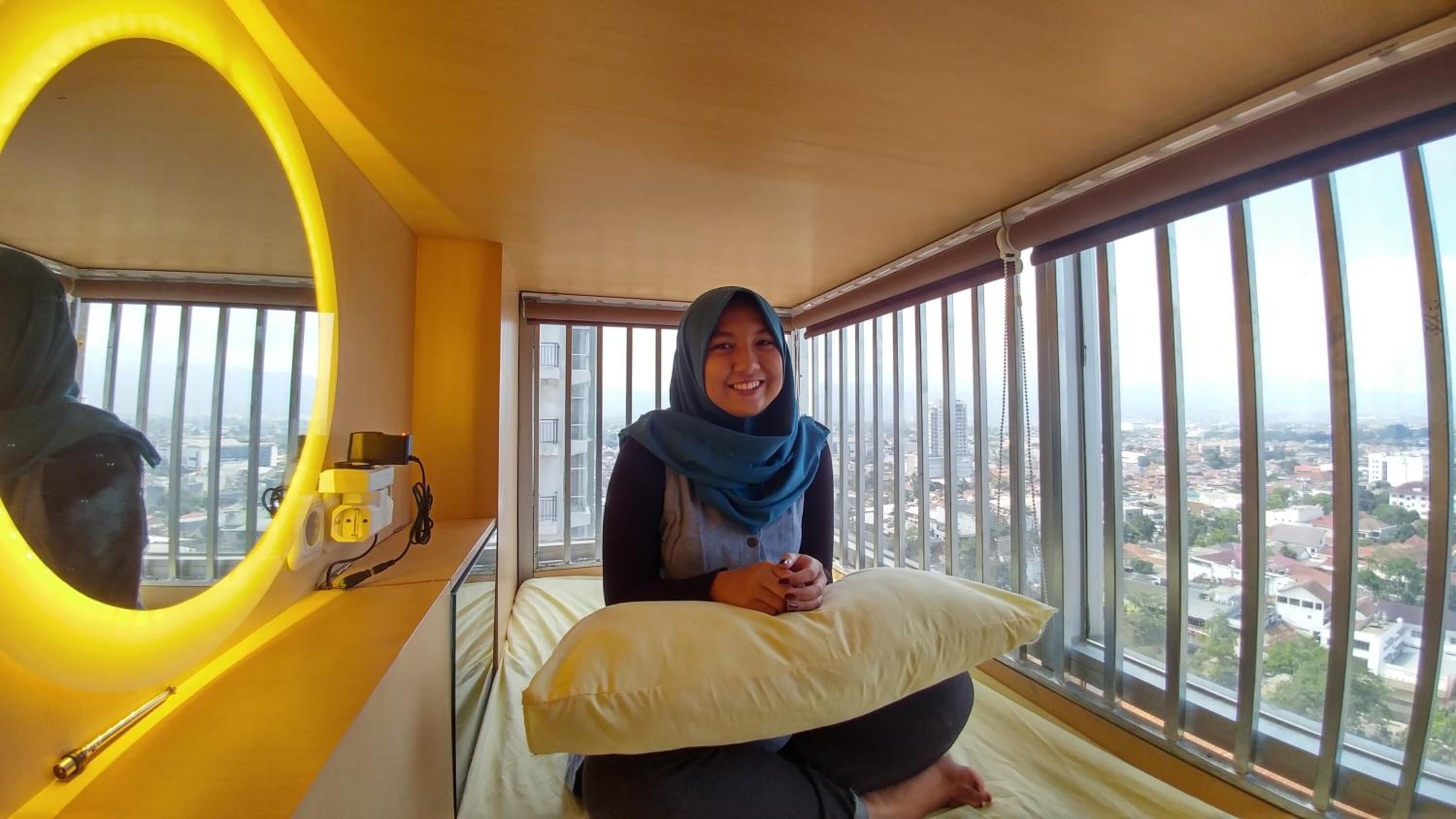 A room with a large window, a bed with a pillow, a mirror, and a wall-mounted device.