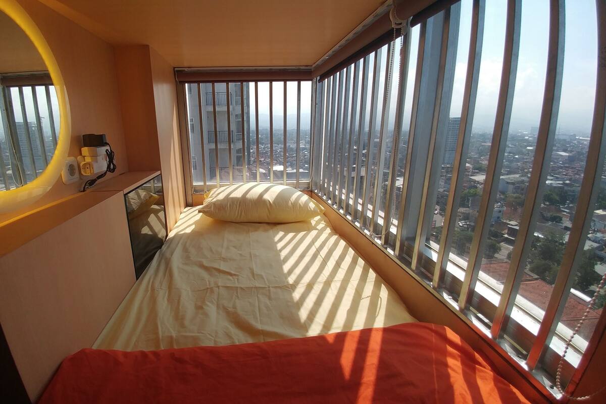 A small bedroom with a bed, a bedside table with a lamp, a window with bars, and a view of the city.