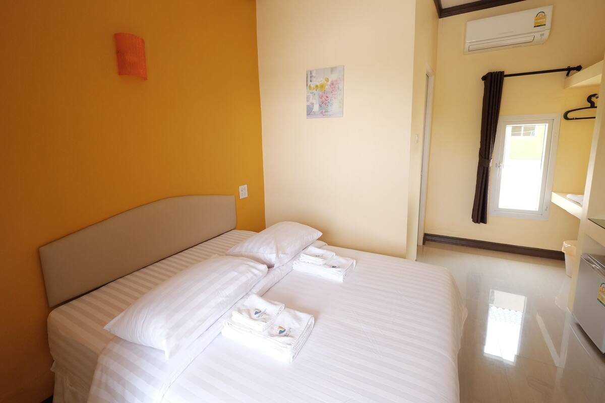 A hotel room with a bed, pillows, a towel, an air conditioner, a window, and a wall-mounted picture.