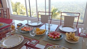 A dining table set with plates, cutlery, and various fruits, with a view of a forested landscape through large windows. - Redhill Kandy (Kandy)