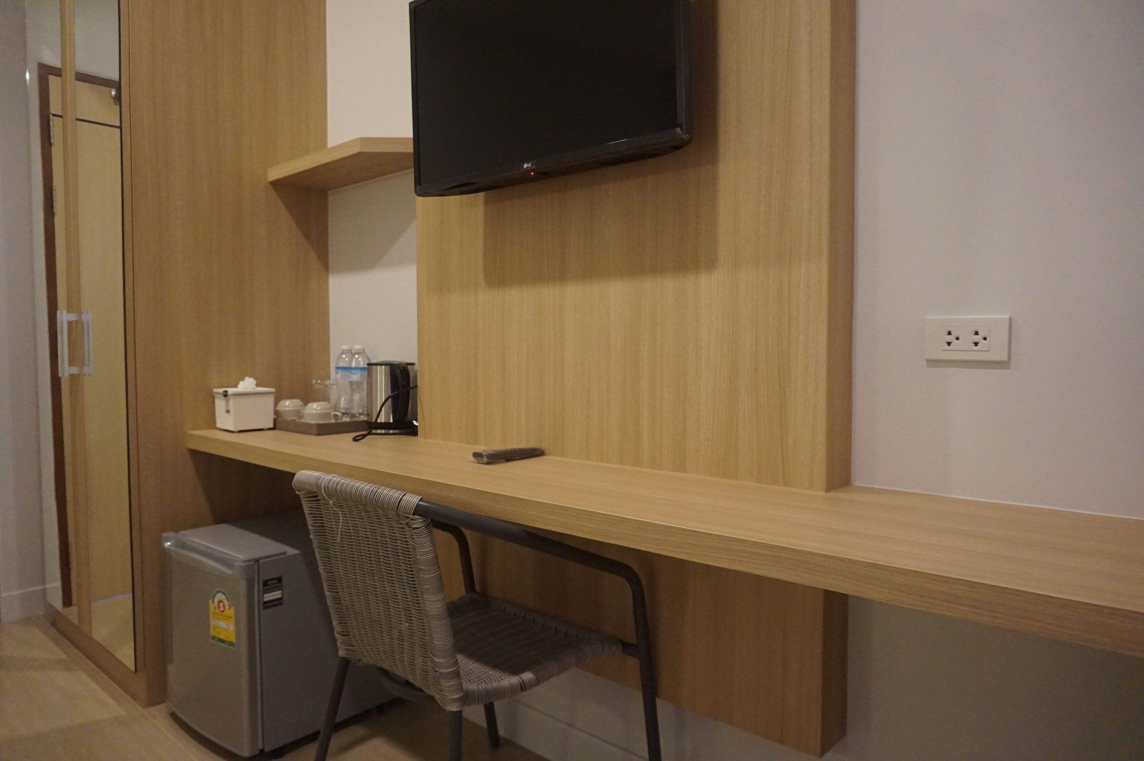 A compact room with a wooden desk, a chair, a small refrigerator, a television mounted on the wall, and a power outlet.