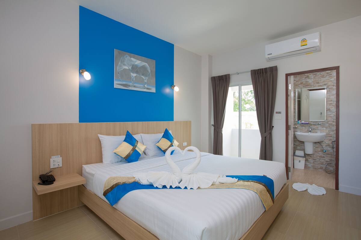 A hotel room with a bed, a bedside table with a phone, a wall-mounted air conditioner, a bathroom with a sink and mirror, and a window with curtains.
