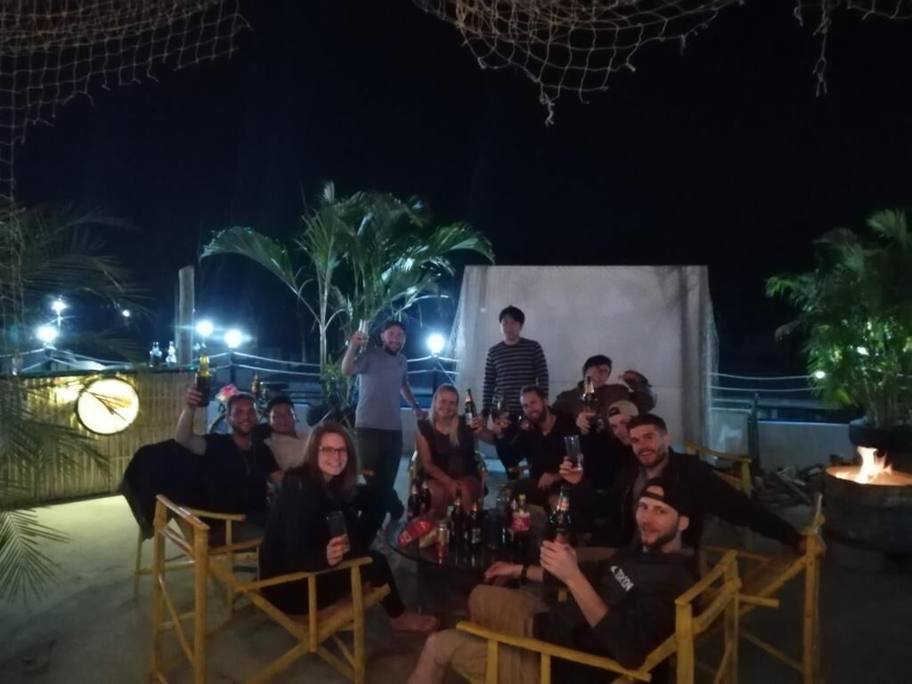Een groep mensen zit aan een buitentafel met drankjes, omringd door palmbomen en een decoratief net erboven.