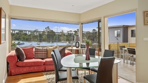 A modern living room with a glass table, leather chairs, and a red sofa. It features large windows with a view of a river and boats. - Manfield City Waterfront View Cottage (Rosny)