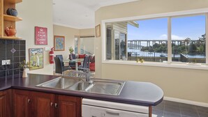 A kitchen with a large window, a sink, and a dishwasher. There is a dining area visible in the background. - Manfield City Waterfront View Cottage (Rosny)