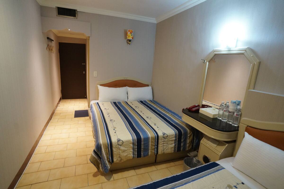 A hotel room with a bed, a bedside table with a mirror, a small desk, and a door leading to another room.