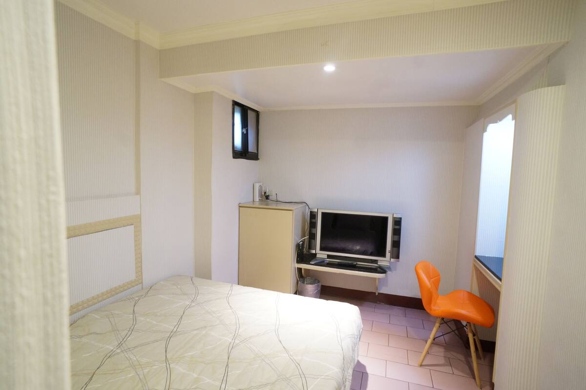A hotel room with a bed, a desk with a computer, a television, and an orange chair.