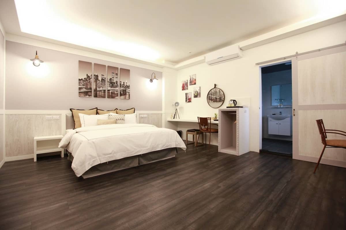 Un dormitorio con una cama grande, un escritorio y una silla. Hay obras de arte en la pared y una puerta que da a otra habitación.