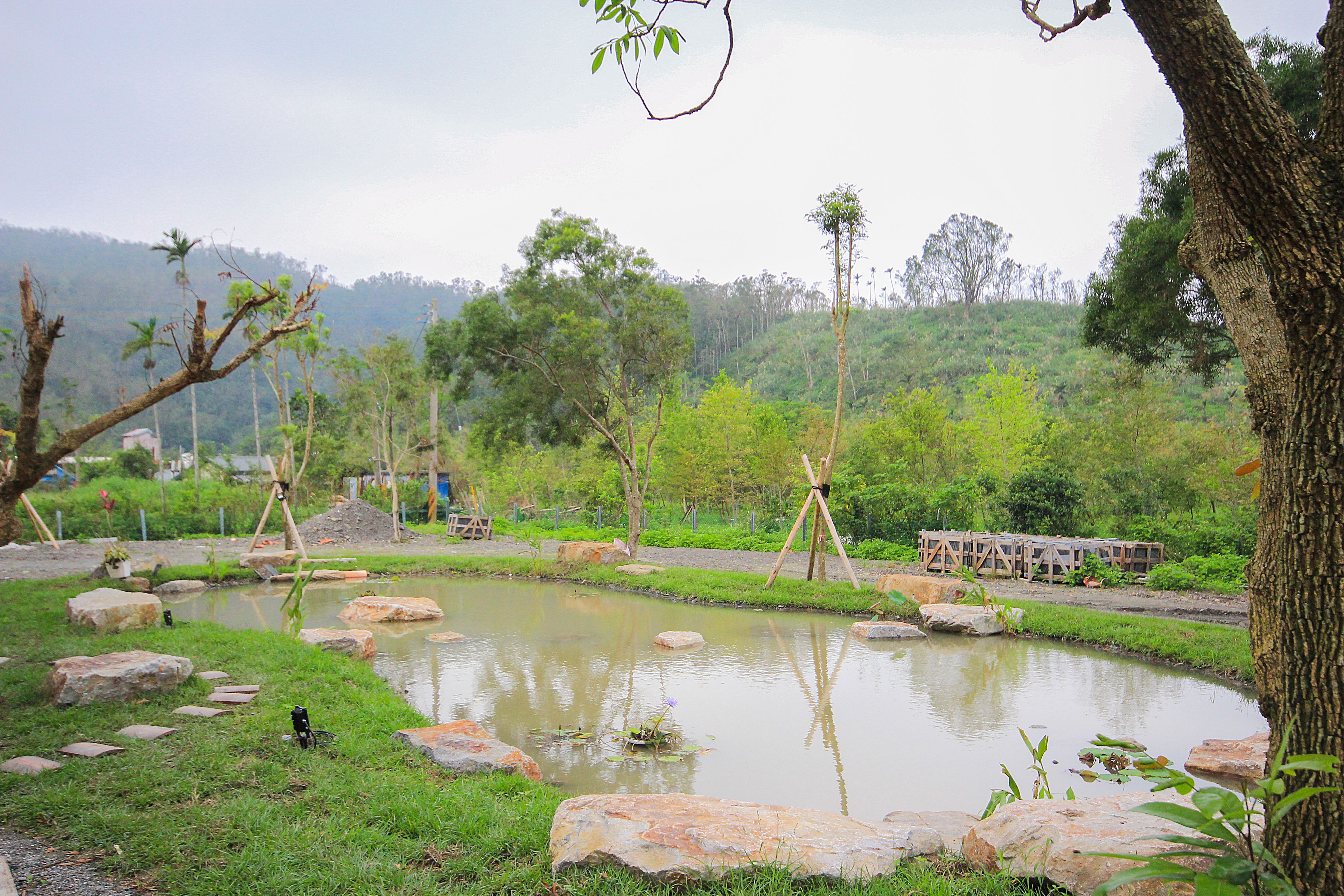 A serene pond surrounded by rocks and greenery, with a wooden bridge and a swing set in the background.