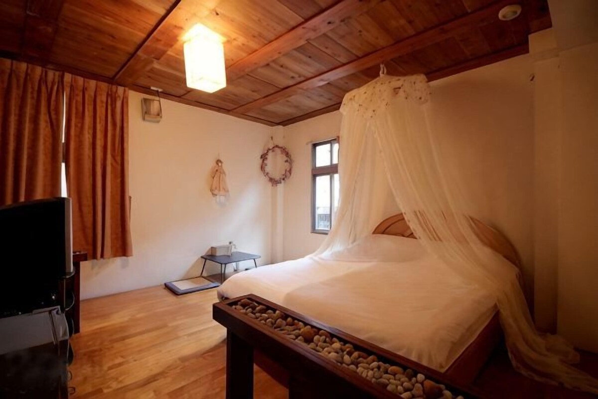 Ein Schlafzimmer mit Holzboden, einem Bett mit Baldachin, einem Nachttisch, einem Fernseher und einem kleinen Tisch mit einem Korb.