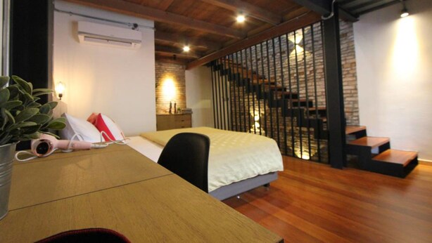 A bedroom with a wooden ceiling, a bed with a white duvet, a desk with a plant, and a staircase with a black railing. - Lorong Jambatan Melaka (Malacca City)
