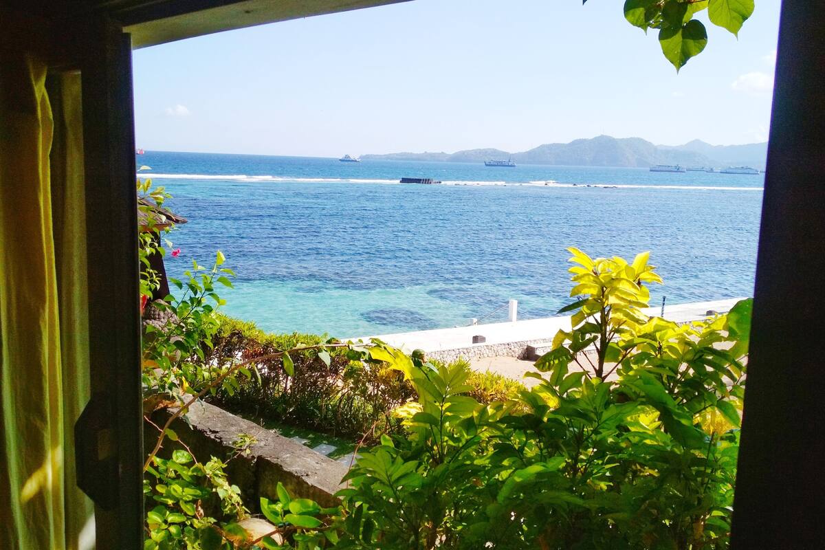 A view through a window of a coastal area with clear blue sea, distant mountains, and greenery.