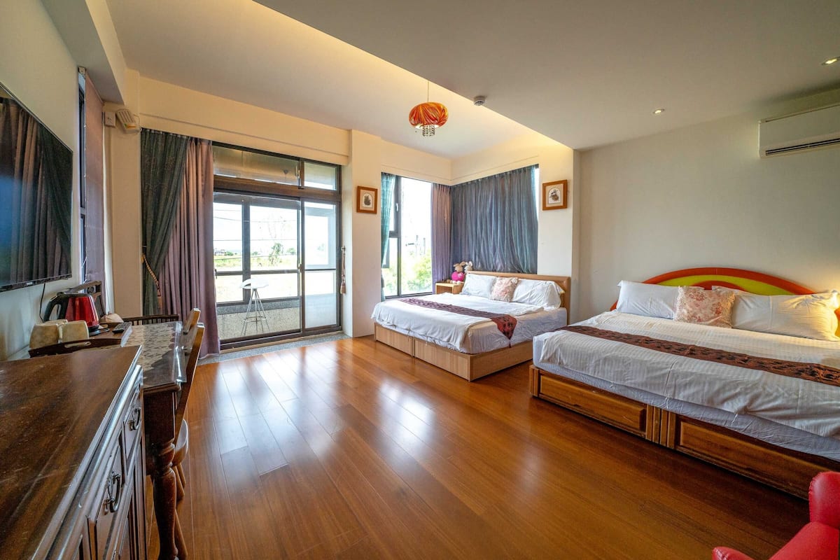 Quarto de hotel com duas camas, uma cômoda de madeira, uma escrivaninha com cadeira e janelas amplas com cortinas.