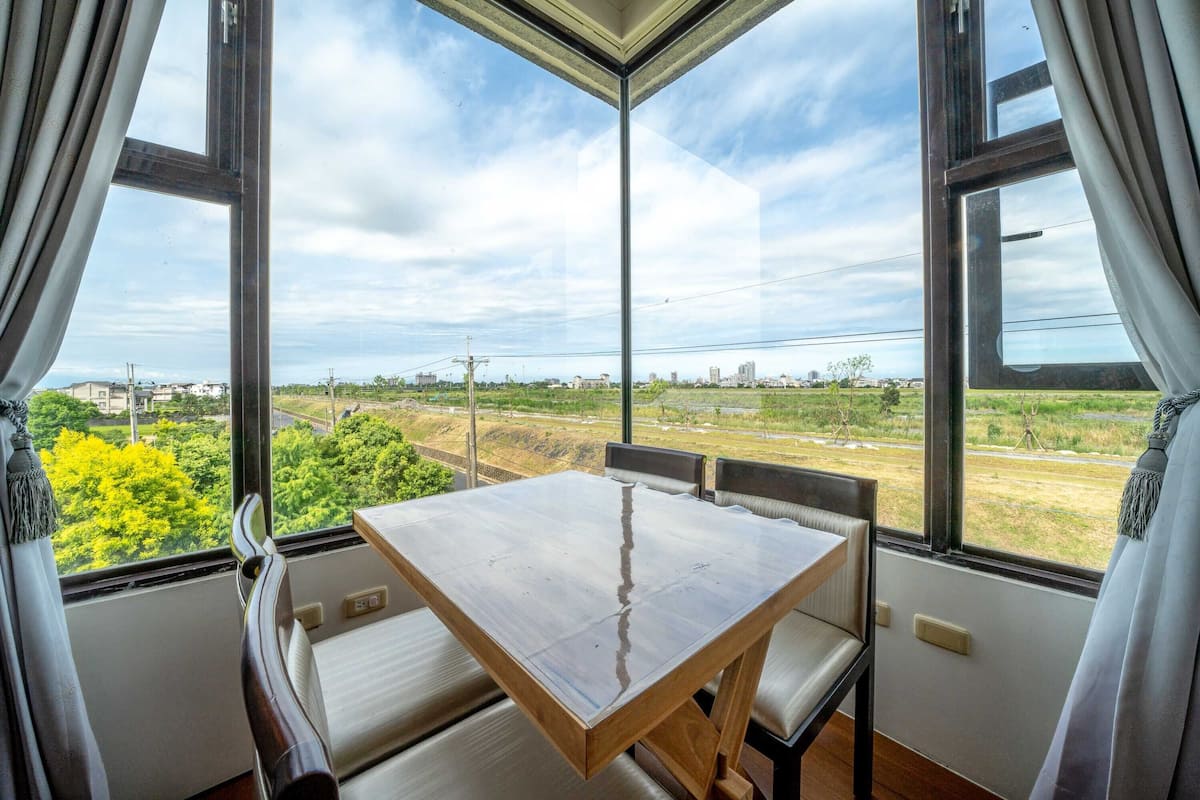 Quarto com mesa, cadeiras, janelas amplas com cortinas e vista para áreas verdes e edifícios.