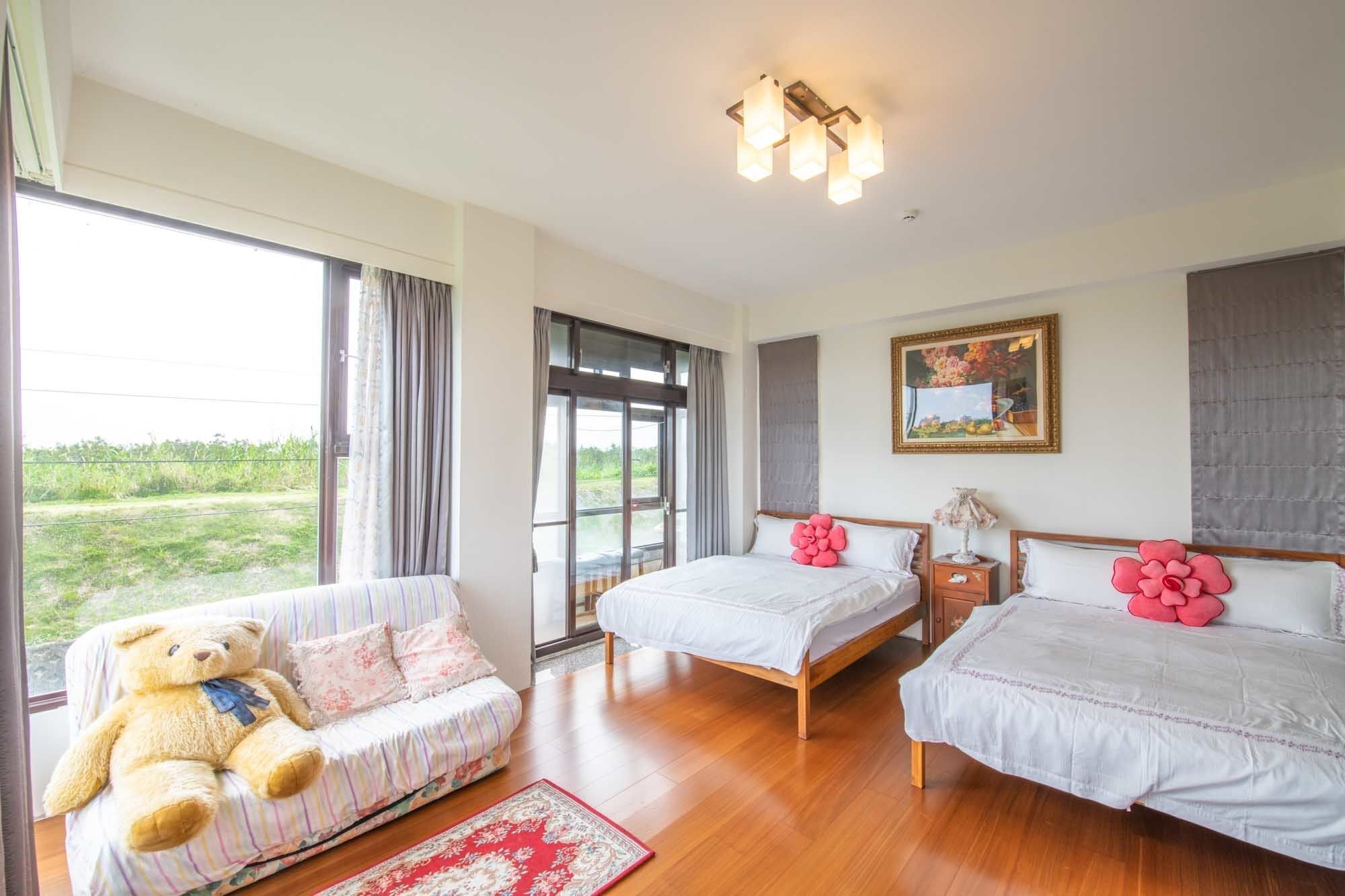 A spacious bedroom with a large window, a sofa with a teddy bear, two beds with pink pillows, and a painting on the wall.