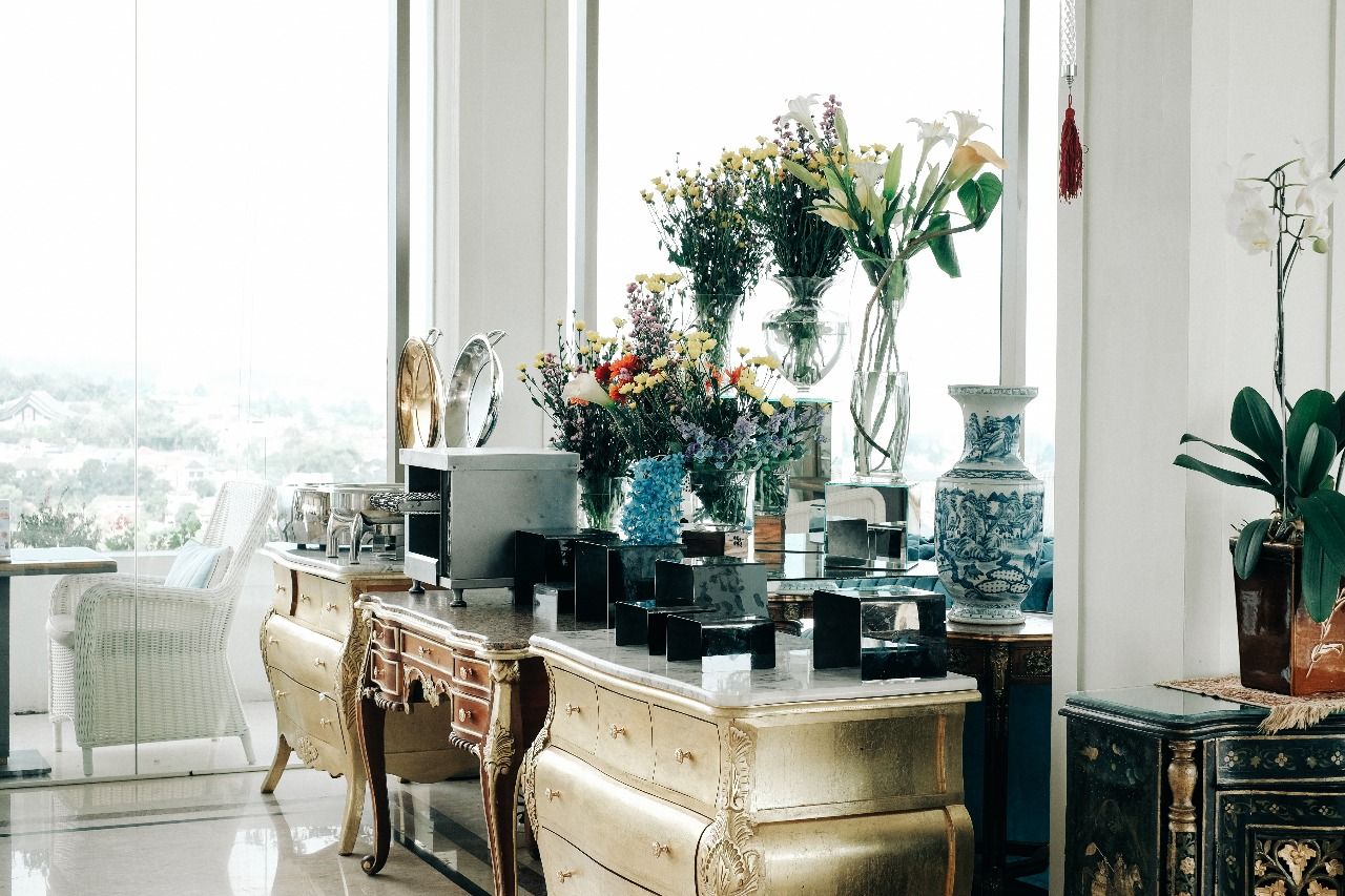 A room with a large window, a white chair, a wooden dresser, and various decorative items including vases and a blue and white ceramic piece.