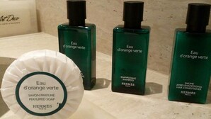 Trois flacons de shampoing et d’après-shampoing verts, un savon blanc en pain et un sac en papier blanc portant les inscriptions « Eau d’orange verte » et « Hermès Paris », le tout posé sur une surface en marbre.