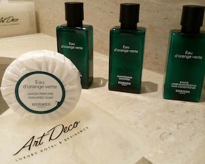 Drie flessen groene shampoo en conditioner, een witte zeep en een witte papieren zak met de tekst 'Eau d'orange verte' en 'Hermes Paris' op een marmeren oppervlak.