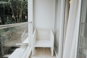 Une chaise en osier blanche sur un balcon doté d’une rambarde en verre, avec un mur blanc et une vue sur les arbres.
