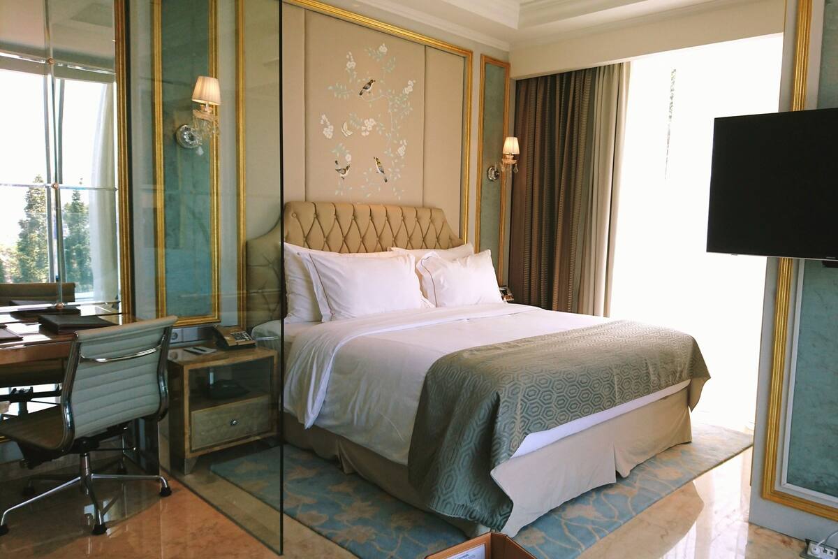 A hotel room with a large bed, a desk with a chair, a television, and a large window with curtains.
