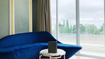 Una habitación con un sofá azul, una mesita redonda con un objeto negro y un ventanal que ofrece vistas a árboles y un estacionamiento.