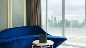 Une pièce avec un canapé bleu, une petite table ronde sur laquelle se trouve un objet noir, et une grande fenêtre donnant sur des arbres et un parking.
