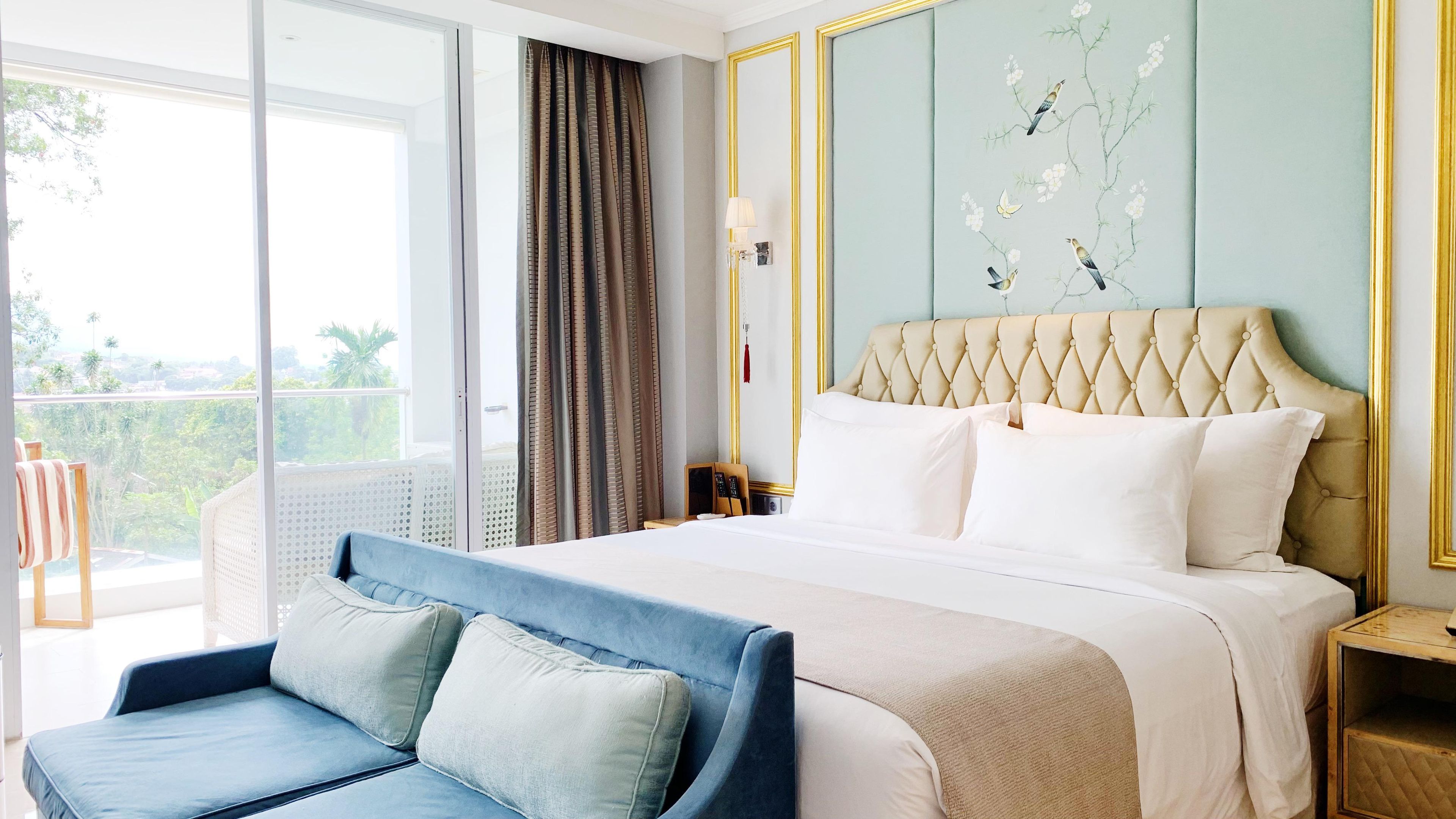 A hotel room with a large bed, a blue sofa, a balcony with a view, and a wall with floral wallpaper.