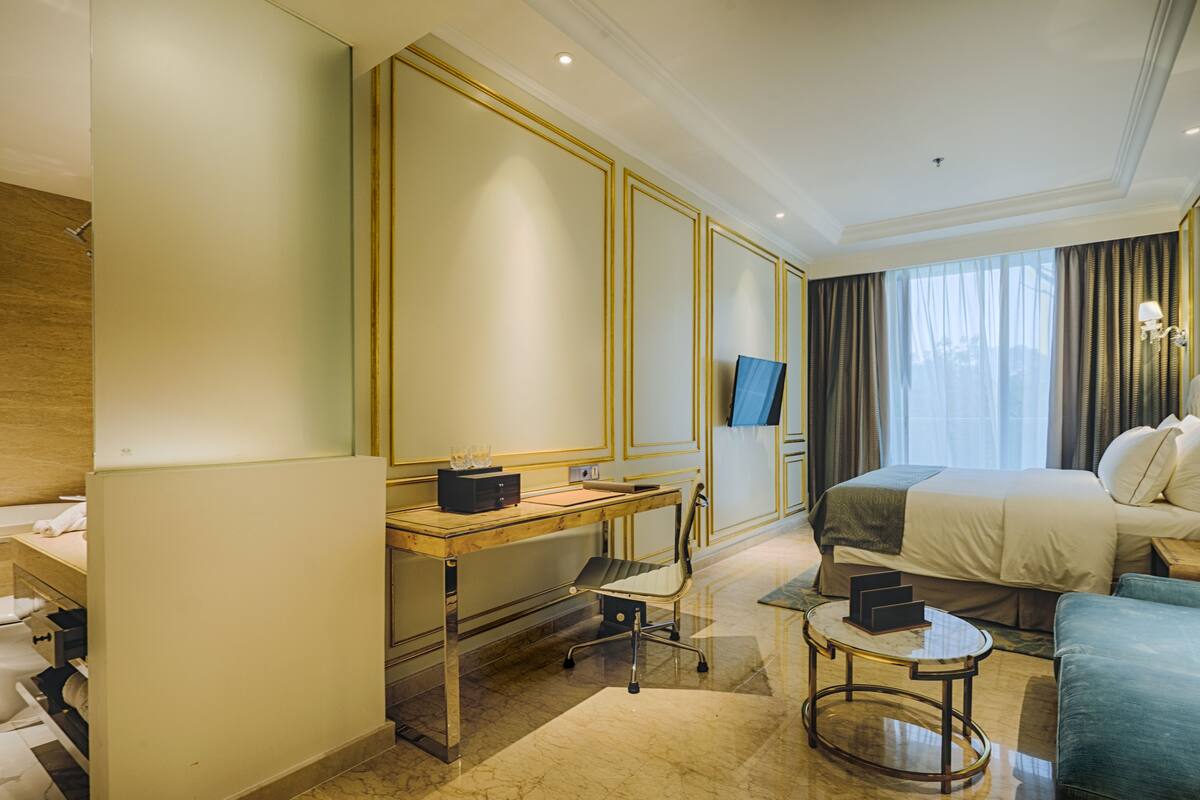 A hotel room with a bed, a desk with a chair, a small round table, a mirror, and a television.