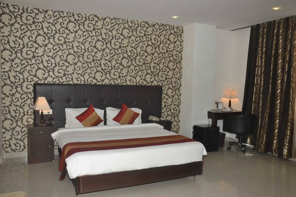A hotel room with a large bed, a nightstand with a lamp, a desk with a chair, and a patterned wallpaper.