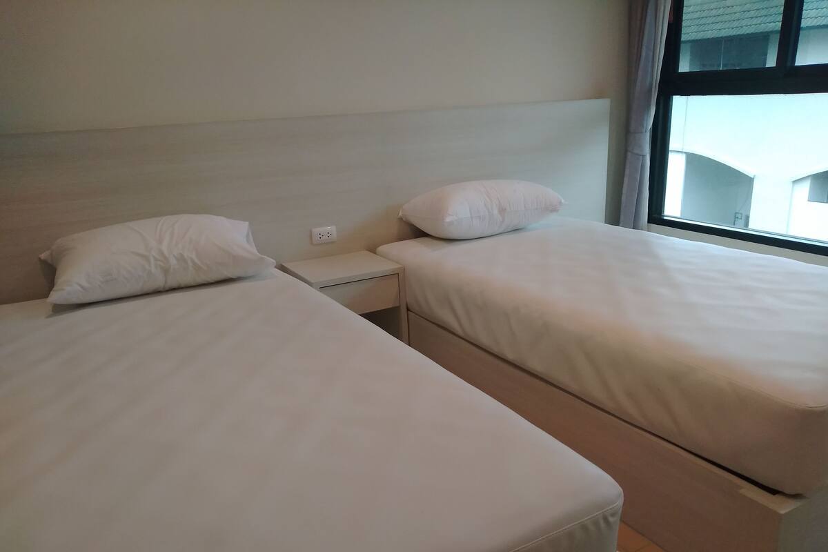 A hotel room with two single beds, a nightstand, and a window with a view of buildings.