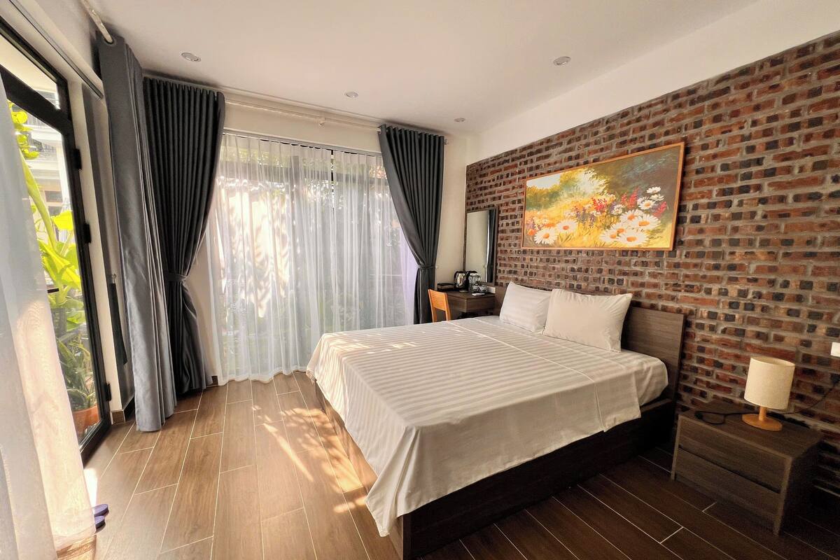 A modern hotel room with a large bed, a brick accent wall, a painting, and a view of greenery through sheer curtains.