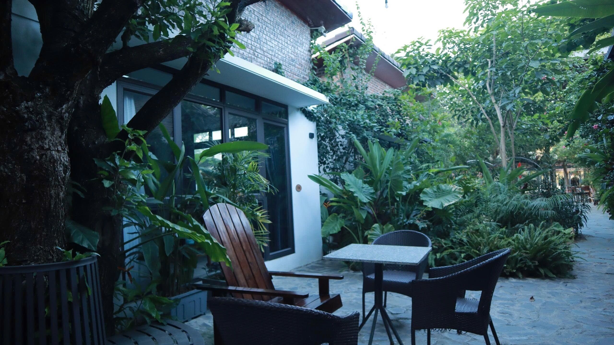 An outdoor patio area with a table and chairs, surrounded by lush greenery and a building with a glass door.