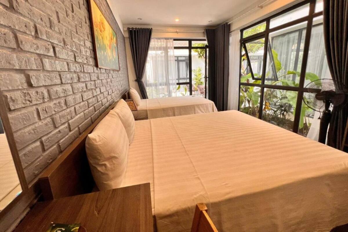 A hotel room with a brick wall, two beds, a wooden table, and large windows with curtains.