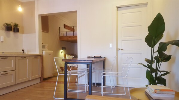 A compact kitchen with white cabinets, a wooden dining table, and chairs. There is a white door and a large green plant in the corner.