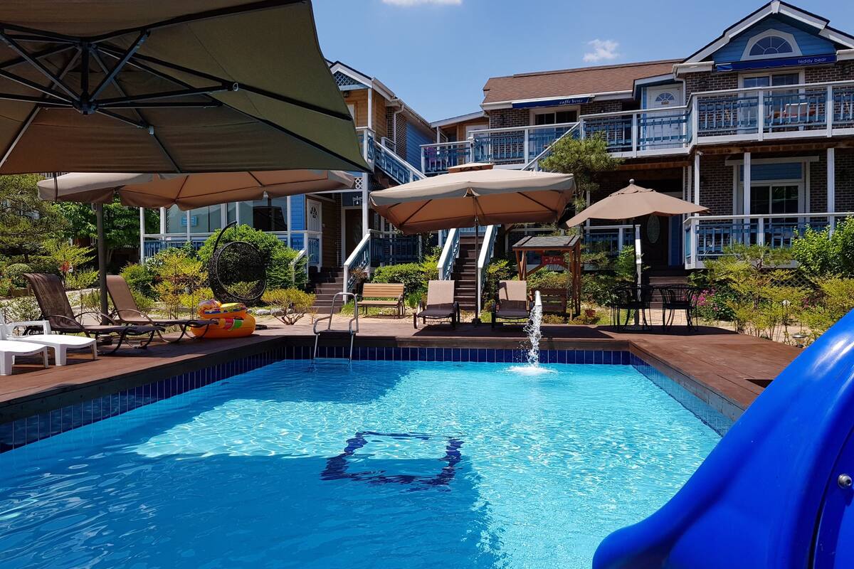 Piscina com escorregador, espreguiçadeiras, guarda-sóis e um edifício com varanda ao fundo.