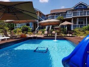 A swimming pool with a slide, lounge chairs, umbrellas, and a building with a balcony in the background.